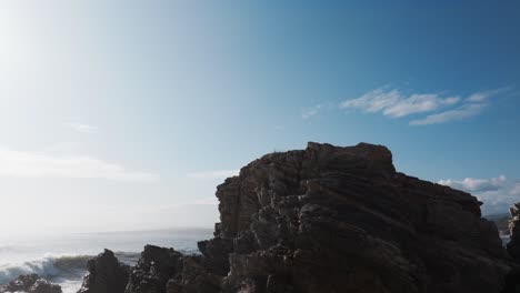 Drone-orbits-around-rock-sheets-protruding-out-from-sandy-beach-as-ocean-waves-crash-into-air