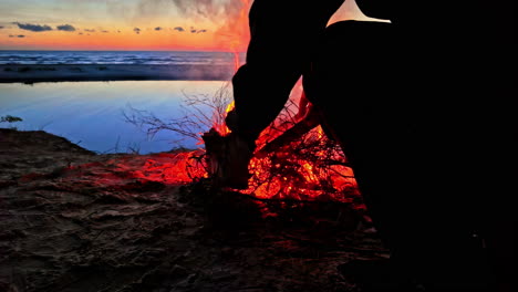 Someone-is-throwing-more-wood-on-a-fire-at-sunset-on-the-beach,-slowmotion-with-copy-space