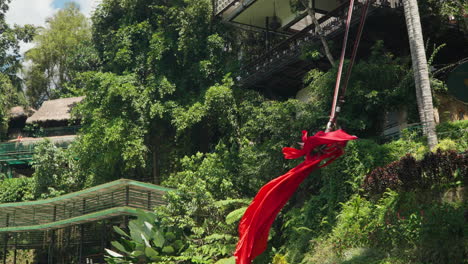 Beautiful-Young-Woman-In-Red-Dress-Swinging-Through-Greenery-In-Alas-Harum-Bali,-Indonesia