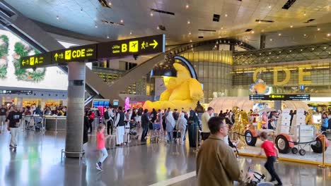 Crowds-of-people-in-terminal-walking-around-big-yellow-teddy-called-untitled-lamp-bear-in-Hamad-International-Airport-in-Doha,-Qatar