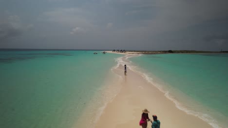 People-walking-on-a-narrow-sandbar-surrounded-by-turquoise-waters-on-a-sunny-day,-aerial-view