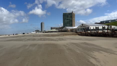 Pov-walk-on-stormy-sandy-beach-of-Netherlands,-Noordwijk-City-with-restaurants-and-hotel