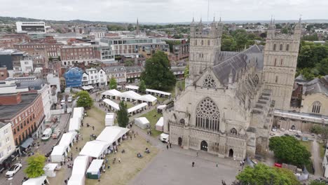 Aerial-view-of-Exeter-Cathedral-with-vibrant-market-stalls-pitched-in-the-grounds,-creating-a-lively-scene-in-Devon,-UK,-July-2024