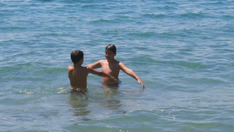 Medium-100fps-slow-motion-footage-of-two-caucasian-brothers-waving-their-hands-in-the-air,-playing-at-the-seashore,-splashing-each-other-with-water