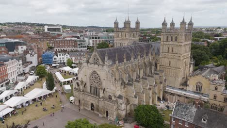 Drone-view-of-Exeter-Cathedral-with-market,-showcasing-a-bustling-scene-of-vendors-and-visitors-in-the-heart-of-Devon,-UK,-July-2024