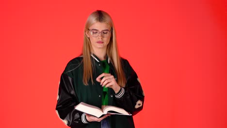 Estudiante-Bastante-Rubia-Haciendo-Un-Gesto-De-Silencio-Mientras-Lee-Un-Libro,-Foto-De-Estudio-Con-Fondo-Rojo