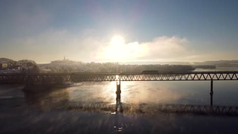 Vista-Aérea-De-Un-Puente-Que-Cruza-El-Río-Glomma-En-Fetsund,-Noruega,-Con-Un-Impresionante-Amanecer-Que-Proyecta-Reflejos-En-El-Agua