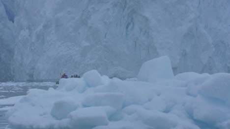 People-in-Boat-Sailing-Under-Massive-Glacier-and-by-Icebergs-on-Sunny-Day-at-Antarctica