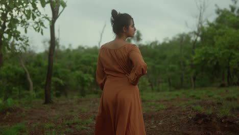 Woman-in-orange-dress-stands-in-a-lush-green-forest,-back-view,-on-a-cloudy-day