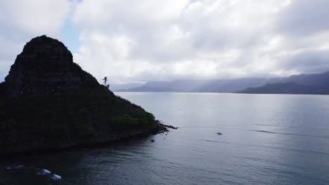 Vista-Aérea-De-La-Isla-Mokoliʻi,-También-Conocida-Como-Sombrero-De-Chino,-La-Silueta-De-La-Isla-Contrasta-Maravillosamente-Con-Las-Aguas-Tranquilas-Y-Reflectantes-Y-Las-Montañas-Brumosas-Al-Fondo.