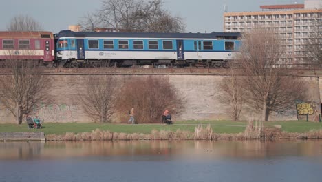 A-sunny-early-spring-day-sees-a-passenger-train-journeying-alongside-a-tranquil-lake,-with-residential-buildings-in-the-background-and-passers-by-strolling-along-the-lake's-banks