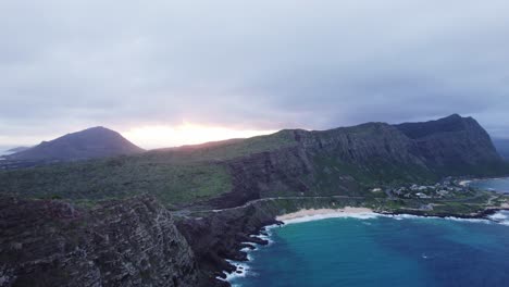 An-aerial-shot-captures-the-breathtaking-view-of-the-sun-setting-behind-the-rugged-cliffs-of-Oahu's-North-Shore,-highlighting-the-stunning-contrast-between-the-lush-green-landscape-and-the-blue-ocean