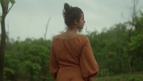 A-woman-in-a-brown-dress-looks-away-in-a-lush-green-forest-setting-on-a-cloudy-day