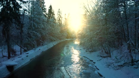 Aerial-dolly-over-river-as-sun-sets-between-trees,-riverbank-leads-to-sunset-glow-of-light-and-snowy-trees