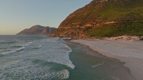 Drohnenaufnahme-Des-Noordhoek-Strandes-Bei-Sonnenuntergang-Mit-Der-Berühmten-Chapmans-Peak-Route-Im-Hintergrund