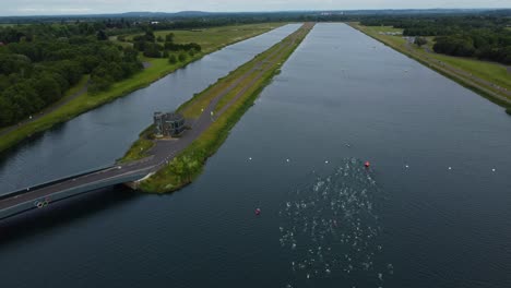 Drone-shot-of-Triathlon-at-Dorney-lake,-triathletes-swimming-towards-the-transition-area-and-cycling-on-the-track