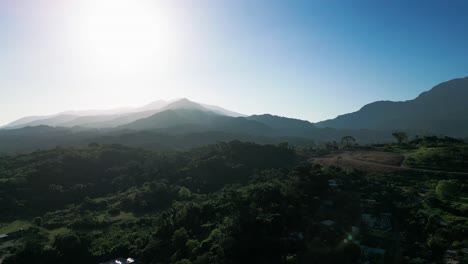 Vista-Aérea-Panorámica-De-Un-Valle-Montañoso-Iluminado-Por-El-Sol,-Destacando-Exuberantes-Bosques-Verdes-Y-Picos-Distantes.