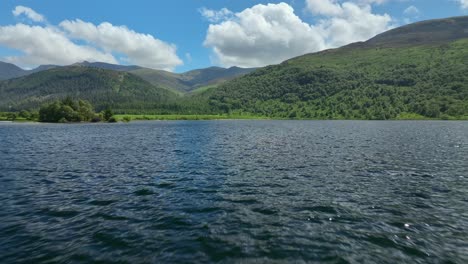 Fast-low-flight-over-blue-lake-waters-towards-forested-hills-and-mountains