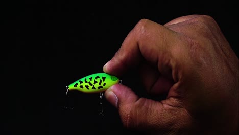 Hand-holding-artificial-lure-of-black-spotted-green-mini-minnow-with-black-background