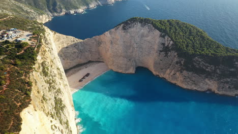 Majestic-aerial-view-of-Navagio-Beach's-Shipwreck-Beach,-surrounded-by-towering-bluffs,-revealing-drone-shot