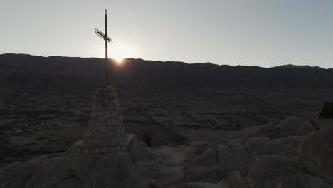 Drone-advancing-and-passing-close-to-the-famous-cross-at-Cerro-de-la-Cruz-in-Tafí-del-Valle,-Argentina,-with-the-sun-rising-behind-the-mountains-at-a-beautiful-sunrise