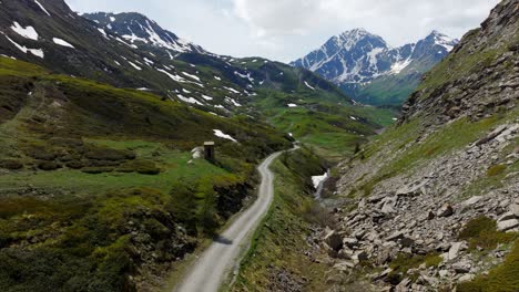 Aerial-View-of-scenic-mountain-road-winds-through-a-lush-green-valley,-flanked-by-rugged,-rocky-hills-and-majestic-snow-capped-peaks-in-the-distance,-under-a-partly-cloudy-sky