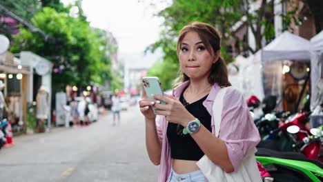 Young-woman-with-light-brown-hair-and-a-pink-shirt,-engrossed-in-her-smartphone-while-standing-on-a-busy-street-lined-with-shops-and-trees