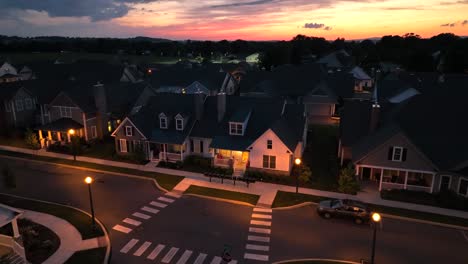 Aerial-dusk-footage-of-a-cozy-suburban-neighborhood-with-charming-houses,-illuminated-streetlights,-and-a-colorful-sunset-in-the-background