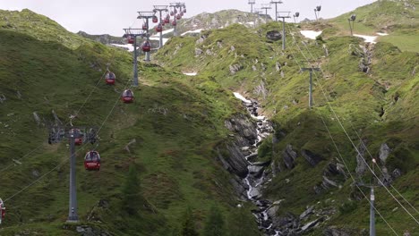 Rote-Seilbahngondeln-Auf-Einem-Grünen-Berg-Mit-Schneeplatten-Und-Einem-Wasserfall-Darunter