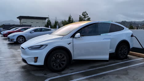 Man-sitting-in-his-white-electric-car,-waiting-for-it-to-be-charged-on-a-high-parking-lot,-pan-shot-in-slow-motion-with-copy-space