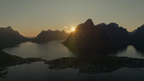 Sun-glowing-behind-silhouette-of-mountains-near-ocean-coastline,-aerial-view