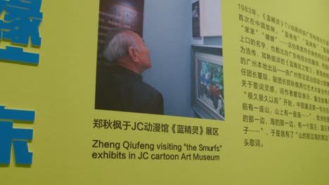 Poster-An-Der-Wand,-Das-Den-Besuch-Von-Zheng-Qiufeng-Und-Qu-Cong-In-Der-Schlümpfe-Ausstellung-Im-JC-Cartoon-Art-Museum-Beschreibt