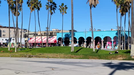 Palmen-Wiegen-Sich-Sanft-Im-Wind,-Während-Besucher-Einen-Gemütlichen-Nachmittagsspaziergang-Entlang-Der-Lebhaften-Strandpromenade-Von-Venice-Beach-Genießen,-Die-Von-Farbenfrohen-Geschäften-Und-Restaurants-Gesäumt-Ist