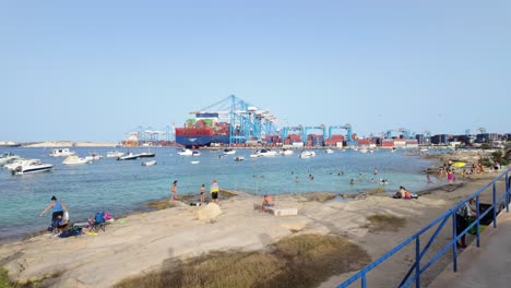 People-swimming-at-Pretty-Bay-in-front-of-the-Maltese-Freeport-with-a-loaded-container-ship-in-Birzebbuga,-Malta