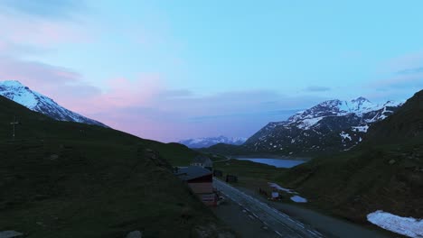 Aerial-view-of-a-winding-road-cutting-through-green-hills-and-valleys,-framed-by-snow-capped-mountains-and-a-tranquil-lake,-all-under-a-vibrant-twilight-sky