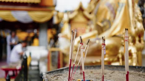 Incense-sticks-burning-in-a-sandy-container,-with-a-golden-temple-background-blurred-out