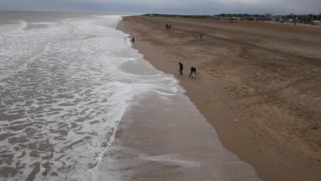 Waves-gently-wash-onto-a-sandy-beach,-where-a-few-people-stroll-and-play-near-the-water's-edge-under-overcast-skies