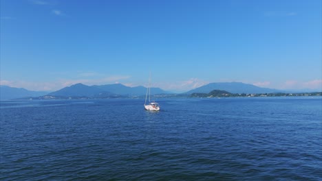 Sonniger-Tag-Luftumlaufbahn-Des-Segelbootes-Auf-Dem-Wasser-Mit-Eingerollten-Segeln,-Lago-Maggiore,-Italien