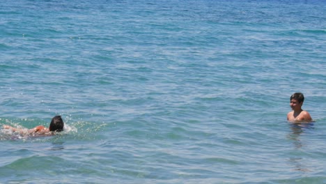 Cámara-Lenta-De-100-Fps,-Toma-Estática-Media-De-Un-Niño-Caucásico-De-8-Años,-Nadando-Hacia-Su-Hermano-De-10-Años,-En-El-Mar-Azul-Cristalino-De-Kalamata,-Peloponeso,-Grecia