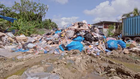 Toma-De-Grúa-De-Un-Vertedero-De-Basura-En-Un-Día-Soleado-Con-Agua-Sucia,-Plásticos-Desperdiciados,-Contenedores-Abandonados,-árboles-Y-Nubes.