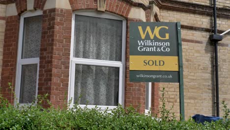 Estate-agent-Sold-sign-outside-red-brick-home-with-green-foliage,-Exeter,-Devon,-UK,-June-2024