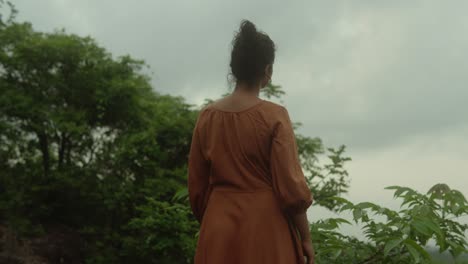 Woman-in-an-orange-dress-stands-in-a-lush-green-forest-under-a-cloudy-sky