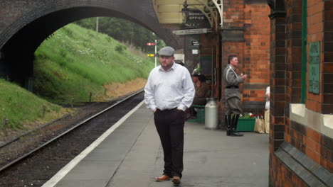 A-man-in-a-flat-cap-walks-along-a-vintage-railway-station-platform-during-the-second-world-war-in-the-channel-islands,-passing-brick-buildings-and-a-booking-office-sign