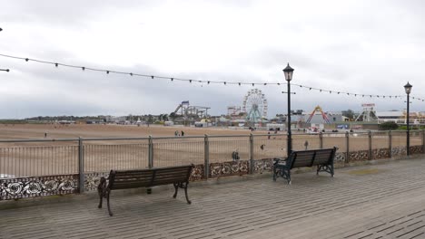 People-walking-on-the-pier-at-the-beach-at-Skegness-with-the-fairground-amusement-park-on-the-shore-at-the-coast