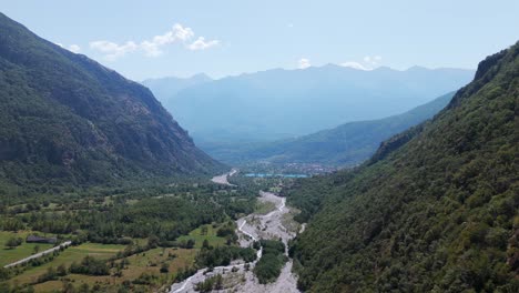 Vista-Aérea-De-Un-Exuberante-Valle-Verde-Ubicado-Entre-Majestuosas-Montañas-Con-Un-Sinuoso-Río-Que-Fluye-A-Través-De-él,-Bajo-Un-Cielo-Azul-Claro