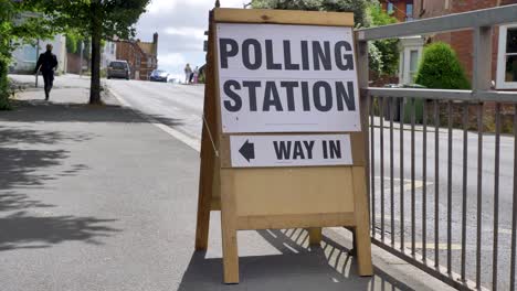 Cartel-De-La-Mesa-Electoral-Con-Gente-Caminando-Para-Votar-En-Un-Día-Soleado,-Exeter,-Devon,-Reino-Unido,-Junio-De-2024