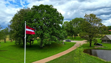 Latvia-flag-waving-near-wooden-hut-log-cabin-by-the-lake-in-nature-field