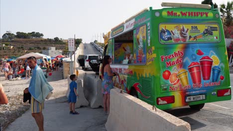 Mama-Kauft-Für-Ihr-Kind-Einen-Frisch-Gemachten-Slush-Von-Einem-Mr.-Whippy-Eiswagen-Am-Strand-Von-Ghar-Ahmar-Bay-Im-Sommer-Auf-Malta