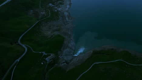 Aerial-shot-of-a-serene-lake-nestled-amidst-towering-snow-capped-mountains,-taken-at-dusk-with-soft-light-reflecting-off-the-water's-surface-and-the-winding-road