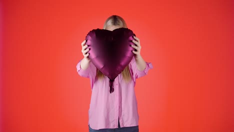 Bonita-Mujer-Rubia-Sosteniendo-Un-Globo-En-Forma-De-Corazón-Púrpura-Contra-Fondo-Rojo,-Foto-De-Estudio